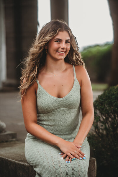 A high school senior in a soft green dress sits outside a grand building, smiling confidently during her senior portrait session. Captured by Nicole Mele Photography in Connecticut, celebrating individuality and graduation milestones.