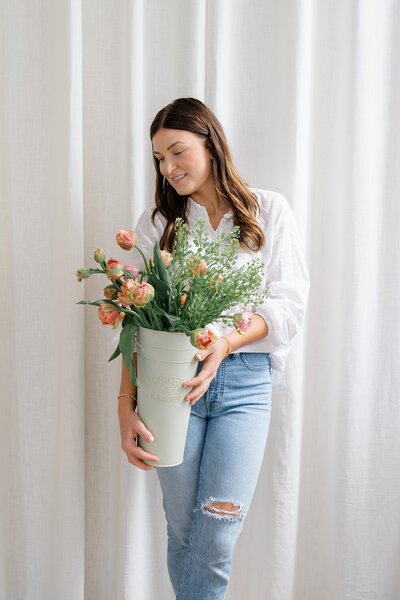 florist brand photos with woman holding a vase of flowers and greenery while standing in front of a curtain and smiling over her shoulder