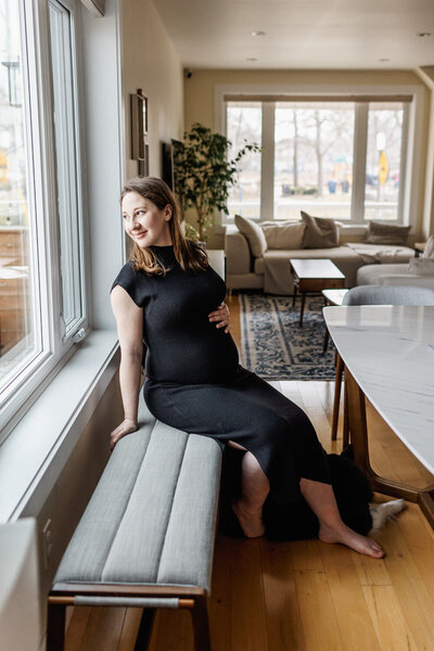Pregnant woman dressed in long black knit dress sitting on a dining bench gazing out the window
