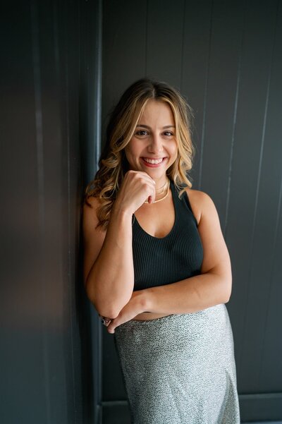 Bee is smiling at the camera with her hand on her hip in front of a yellow background wearing a casual outfit of blue jeans and a white tank top