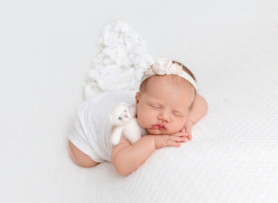 Newborn baby girl sleeping on her belly with her hands resting under her chin. Baby is draped in a white knit wrap with a tiny white teddy resting under her arm. Captured by Rochel Konik top Brooklyn and NYC Newborn Photographer.