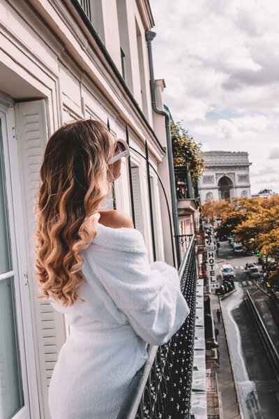 A person with long, wavy blonde hair in a white robe stands on a balcony, gazing at the Arc de Triomphe in the distance. The scene includes a city street lined with autumn-colored trees and buildings.