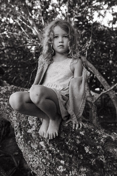 Little girl with wild curls sitting in a tree looking out , she is barefoot and her jumper is falling off of her