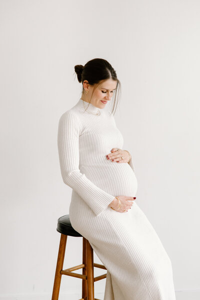 Expecting mother sitting on stool and looking toward bump during studio maternity photos in Indianapolis