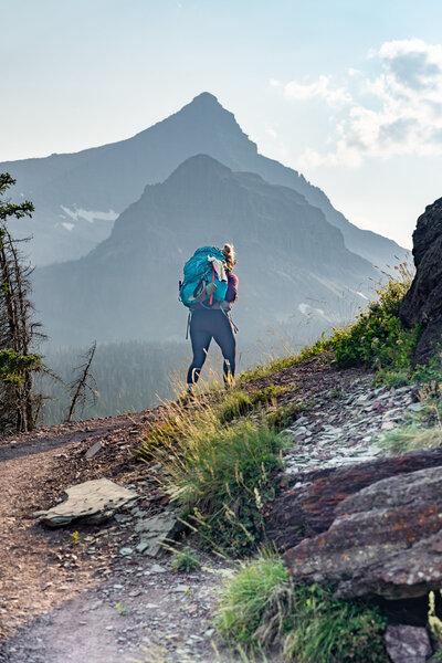 A female backpacker rounds the corner on a trail with Rising Wolf mountain behind her in Glacier National Park in Montana
