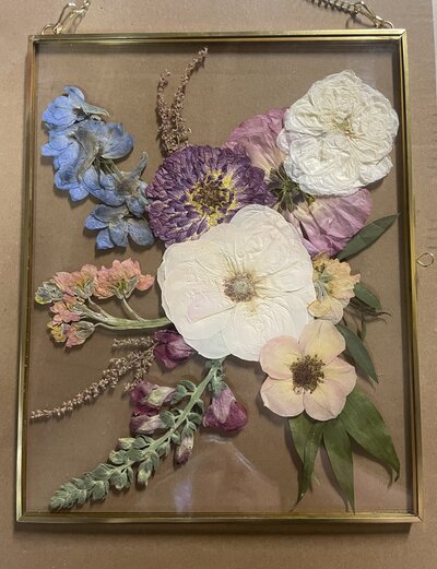 Romantic pressed flower keepsake showcasing white roses, purple zinnias, and delicate greenery in a glass gold frame, ideal for wedding bouquet preservation