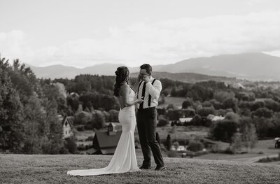 A couple twirls gracefully on a grassy hill, captured by an elopement photography New England expert. The woman in a flowing dress and the man in suspenders overlook scenic valleys with trees and houses, against rolling hills under a cloudy sky. The timeless black and white image captures their love.