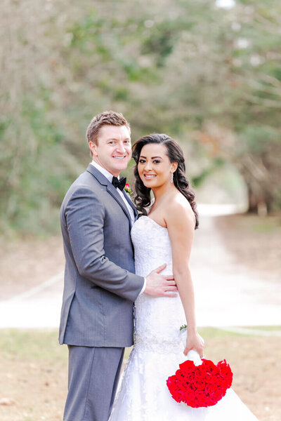 A bride and groom hug and smile while standing in a park holding a bright red bouquet