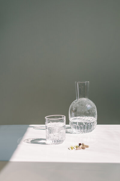 Clear glass pitcher and cup with vitamins on a sunlit surface for hydration and wellness. 