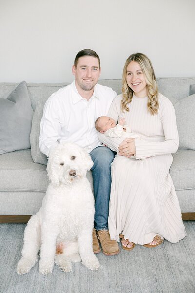 neutral toned newborn session of a mom and dad hold their baby while sitting on a couch with their dog sitting on the floor next to them