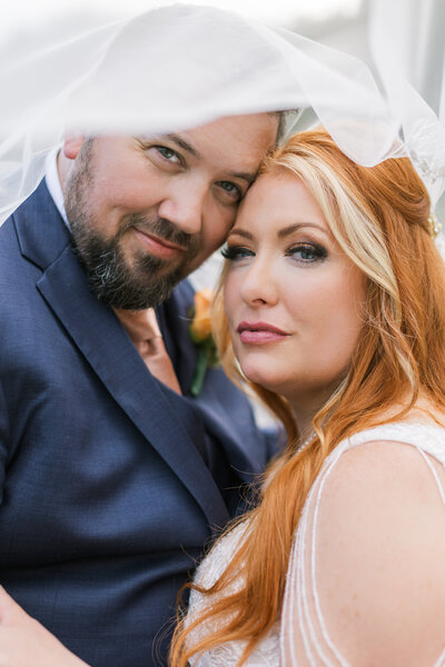 A bride and groom in a classic pose looking at the camera as the veil is over their heads. 