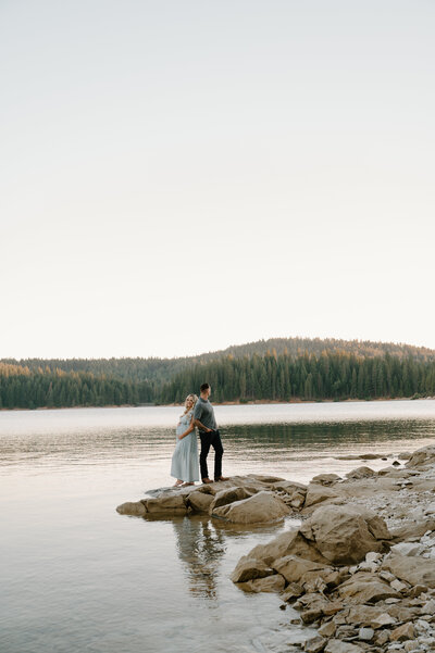 Expecting parents’ hands resting on pregnant belly during an outdoor maternity shoot in the mountains near a lake