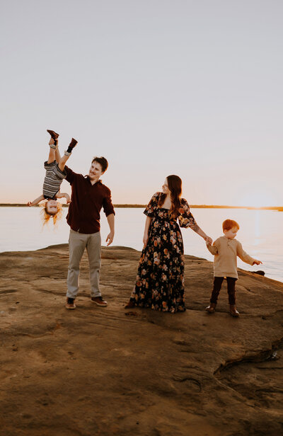 A dad holding his toddler upside down while the mother looks on with a laugh and holds hands with her son at Grapevine Lake in Grapevine, TX
