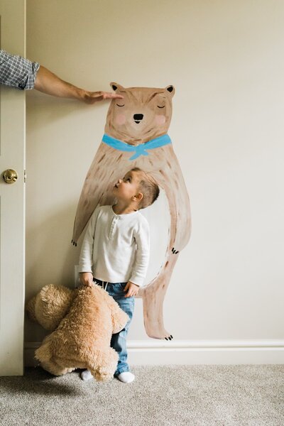 A child looks up at a large bear illustration on the wall while holding a teddy bear, with an adult's hand reaching out towards the illustration, captured by a skilled photographer in Pittsburgh.