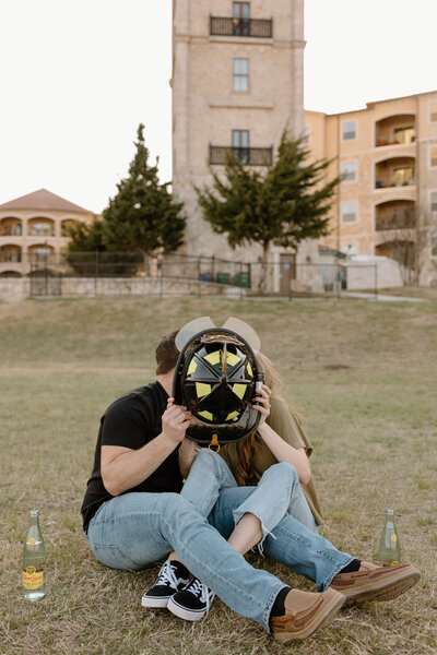 man and woman cuddling on the grass with firefighter helmet in front of their heads and kissing