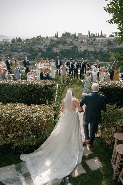 Bride and her bridesmaids walking through the village of Gordes, France in mismatched white lace dresses for a summer wedding