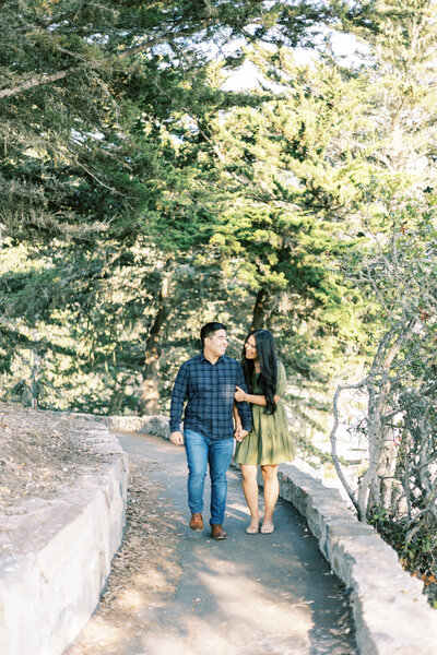 couple holding hands walking along dirt path lined with coastal trees
