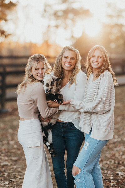 Three women posed and holding a goat surrounded by fall landscape.