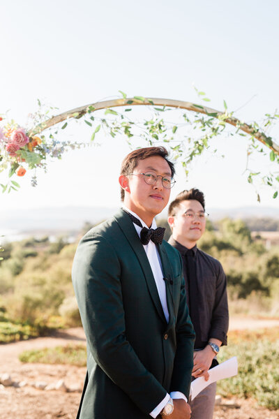 A portrait of the groom awaiting his bride as he stands in front of the arch with the officiant behind him. The floral design by Moonlight Floral Co allows the guests to still enjoy the scenic views of Dos Pueblos Orchid Farm in Santa Barbara.
