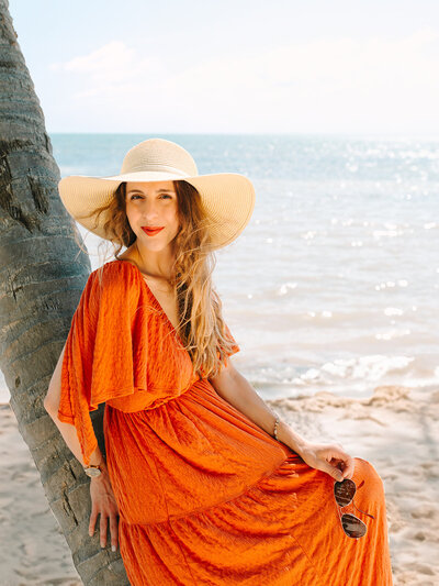 Kilee in an orange dress and straw hat sitting on a palm tree front of the ocean