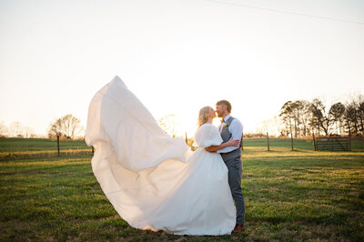 Newlyweds nuzzle each other in front of the golden sunset while her wedding gown flows in the wind