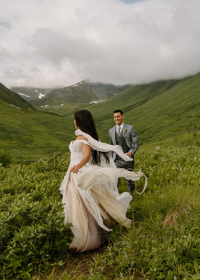 Bride’s flowing gown caught in the wind as she and the groom explore the wild green valleys of Hatcher Pass, Alaska during their adventurous elopement.
