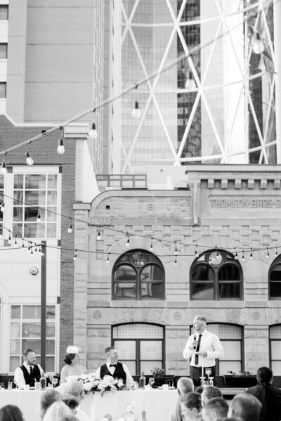 Black and white photo of the best man giving a speech during a rooftop wedding reception at the Hyatt Hotel in downtown Calgary with guests listening and city views in the background
