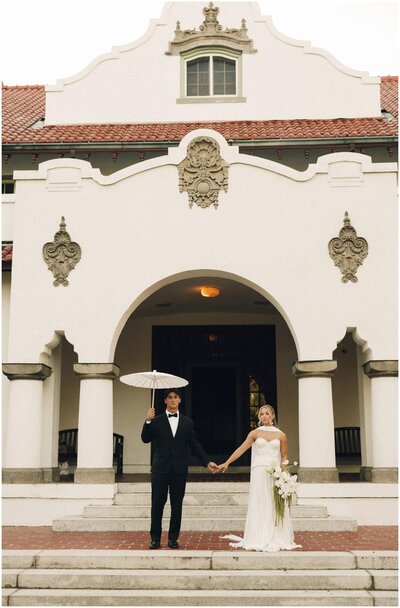 Newlyweds hold hands, an umbrella and large white bouquet on the front steps of their venue