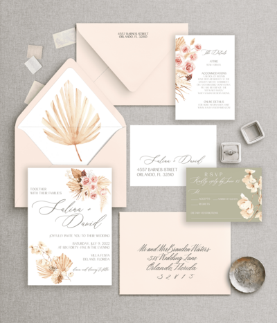 Flat lay of a peach, white, and sage-toned wedding invitation suite with floral accents
