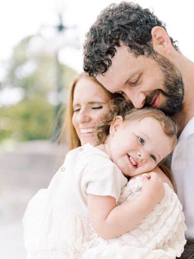 Toddler girl happily snuggles into her mother's shoulder while father snuggles them both