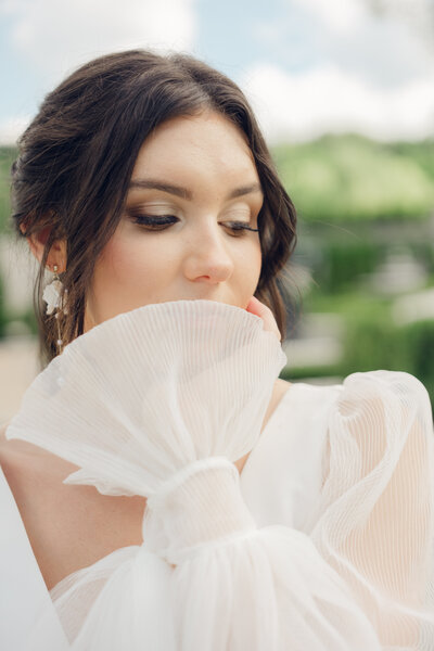 Bride in sheer, pleated gown sleeve looking down with a serene expression outdoors.