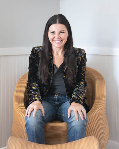 branding session portrait of beautiful business owner with long dark hair in a black patterned jacket and jeans sitting on interesting wicker chair