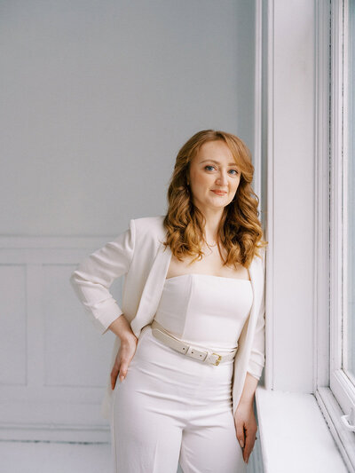 A person with long, wavy hair stands beside a window, exuding elegance in a white outfit consisting of a blazer, top, and pants. The poised demeanor suggests they might be a wedding planner in Calgary.