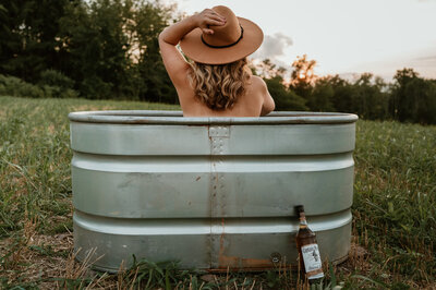A woman with her back to the camera sitting in a watering trough with only a hat on. 