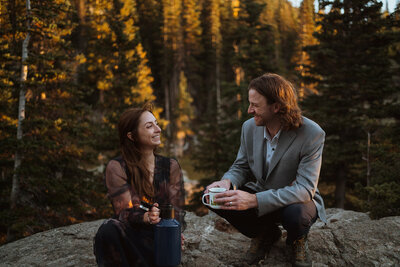 the bride and groom are drinking coffee as the sun rises. the bride is holding a navy blue canteen and the groom is holding a coffee cup with pine trees on it. they are sitting with yellow aspens and pine trees in the background. they are both smiling at each other in wedding attire.