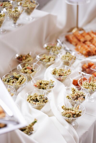 Individual servings of pasta salad in clear cups with mini forks, arranged on white fabric, with another appetizer visible in the background.