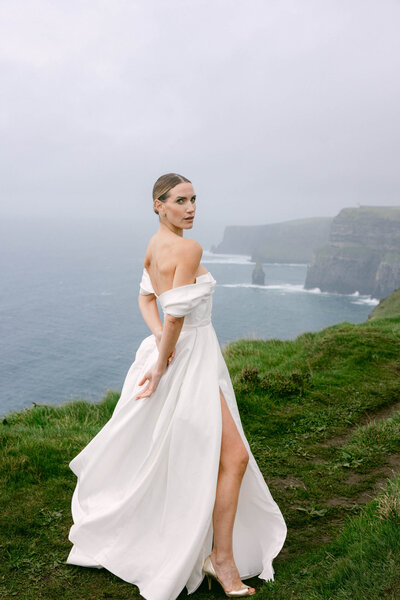A bride in a powerful pose with her hands behind her back looking at the camera during her bridal shoot on the Cliffs of Moher in Ireland