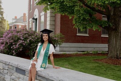 A smiling graduate in a white dress and green stole sits on a stone ledge, her graduation cap proudly perched. Surrounded by blooming bushes and trees, the scene evokes the charm of New England's historic brick buildings with lush greenery cascading around her.