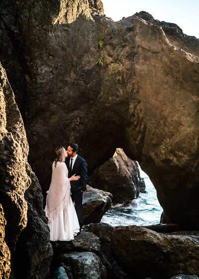 a couple in their wedding attire snuggle in the crevice of a sea stack. through the hole in the rock, we glimpse the ocean waves rolling behind them during their adventure elopement 