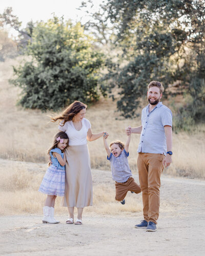 Family of four with parents and two children playfully pose during San Jose photoshoot, with the parents swinging the toddler boy 