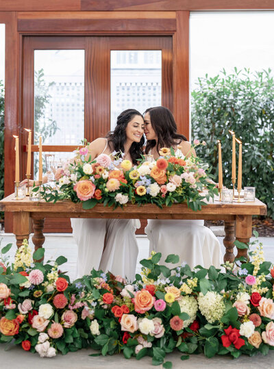 Two women in white dresses sit at a wooden table adorned with vibrant flowers and candle holders. They share a joyful moment, surrounded by floral arrangements of roses and other blooms. Large windows and greenery are in the background.