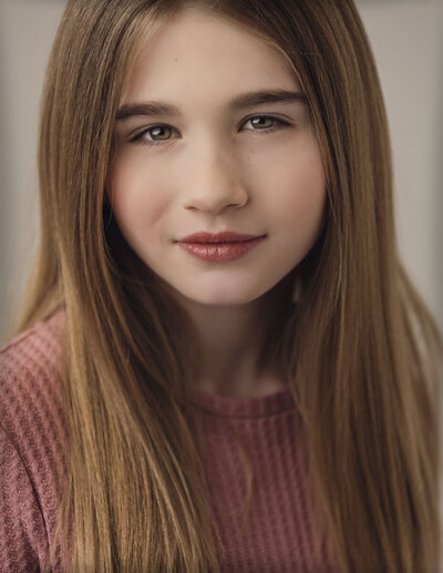 A young person with straight, long brown hair wears a textured pink top. They look directly at the camera with a calm, neutral expression.