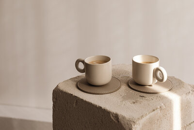Two artisanal beige coffee cups sit on matching leather coasters atop a textured concrete pedestal. The soft, monochromatic palette enhances the cozy, sophisticated aesthetic. Styled for a local café’s social media, this image highlights a warm and inviting coffee experience.