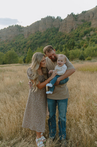 A husband and wife hold their 1 year old baby in their arms as they smile and look at her. Behind them is a beautiful landscape.