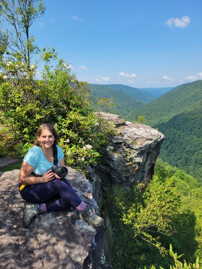 Joy Granados sitting with her camera on a cliff overlooking mountain views, adventurous behind-the-scenes image of Capture Joy Photography, Northern Virginia family photographer.
