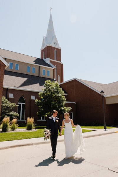 A newlywed couple holding hands and walking outside a church