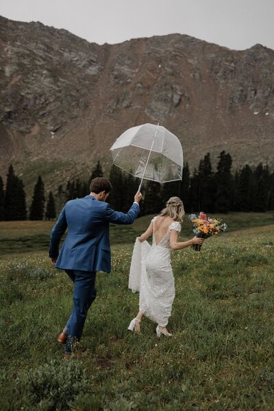 Bride and groom walk through alpine field with umbrella during rainy mountain elopement.