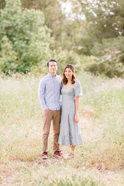 Kohnur Bride with soft romantic makeup and loose curls stands beside her groom in a sunlit garden field, capturing a serene and timeless engagement portrait with effortless wedding elegance