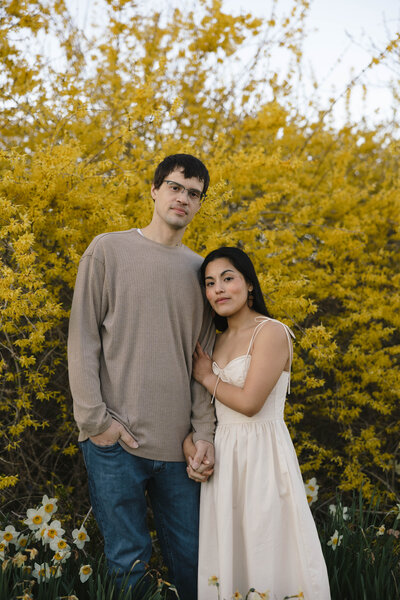Corey embracing Gaby in front of a yellow blooming bush