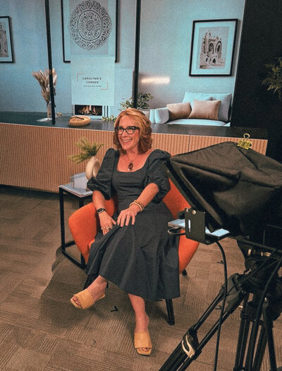 Carolynn Santaniello seated in a studio setting, smiling during a video shoot with camera equipment in the foreground.